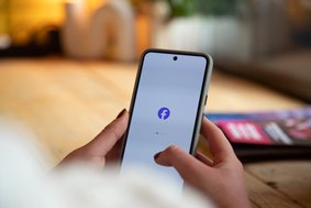 Hände mit Smartphone in der Hand. In der Mitte das Facebook Logo.