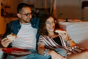 Mann und Frau sitzen auf dem Sofa, essen Pizza und schauen etwas.